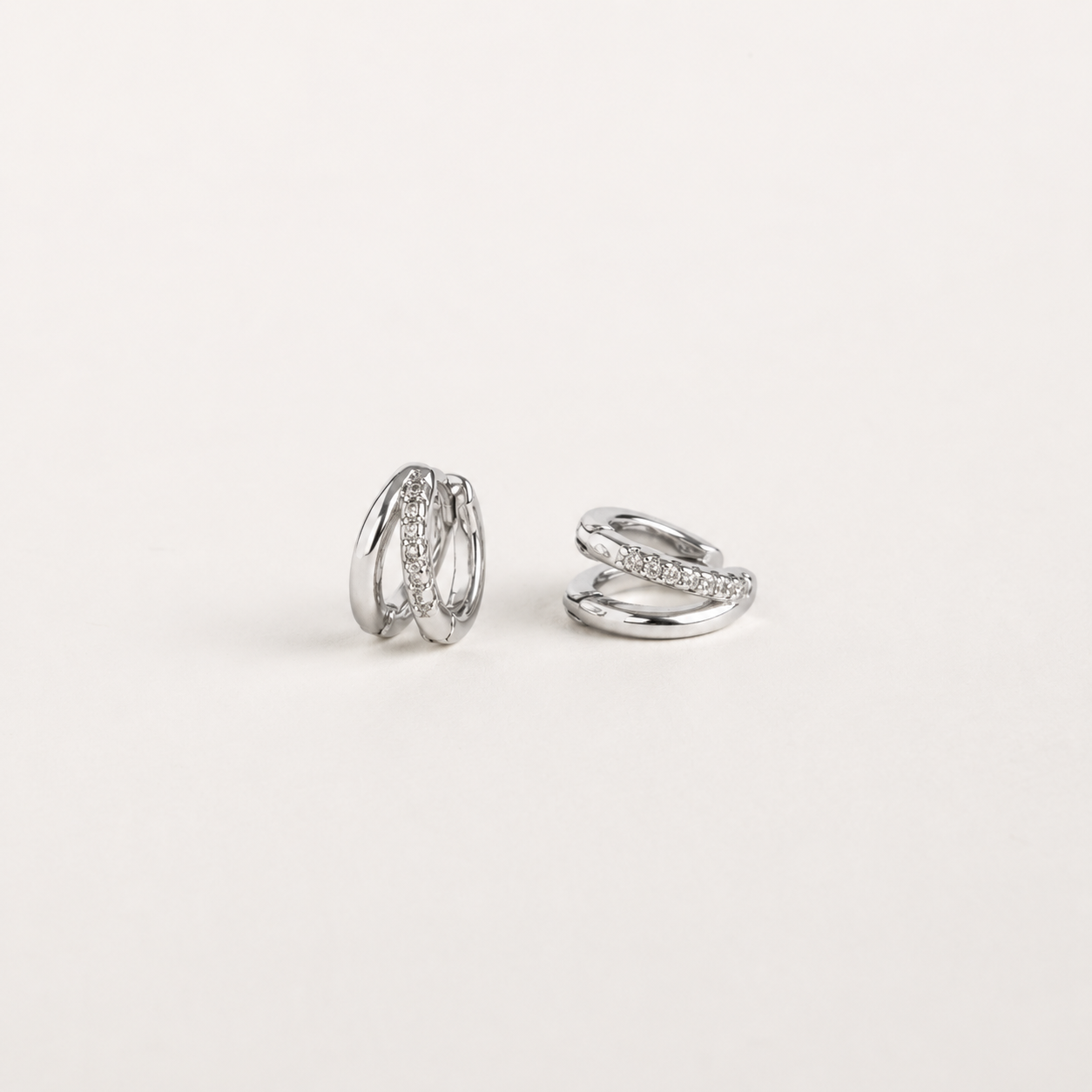 A pair of silver-toned huggie hoop earrings with a twisted, layered design, one earring featuring a row of small clear stones, photographed on a clean, light off-white background.